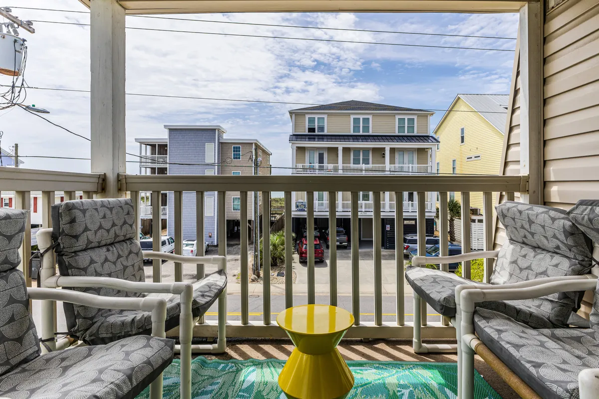 An inviting exterior shot featuring two armchairs and a yellow side table on a balcony, overlooking neighboring buildings. The setup suggests a relaxed spot to unwind after beach activities and enjoy the coastal neighborhood vibe.