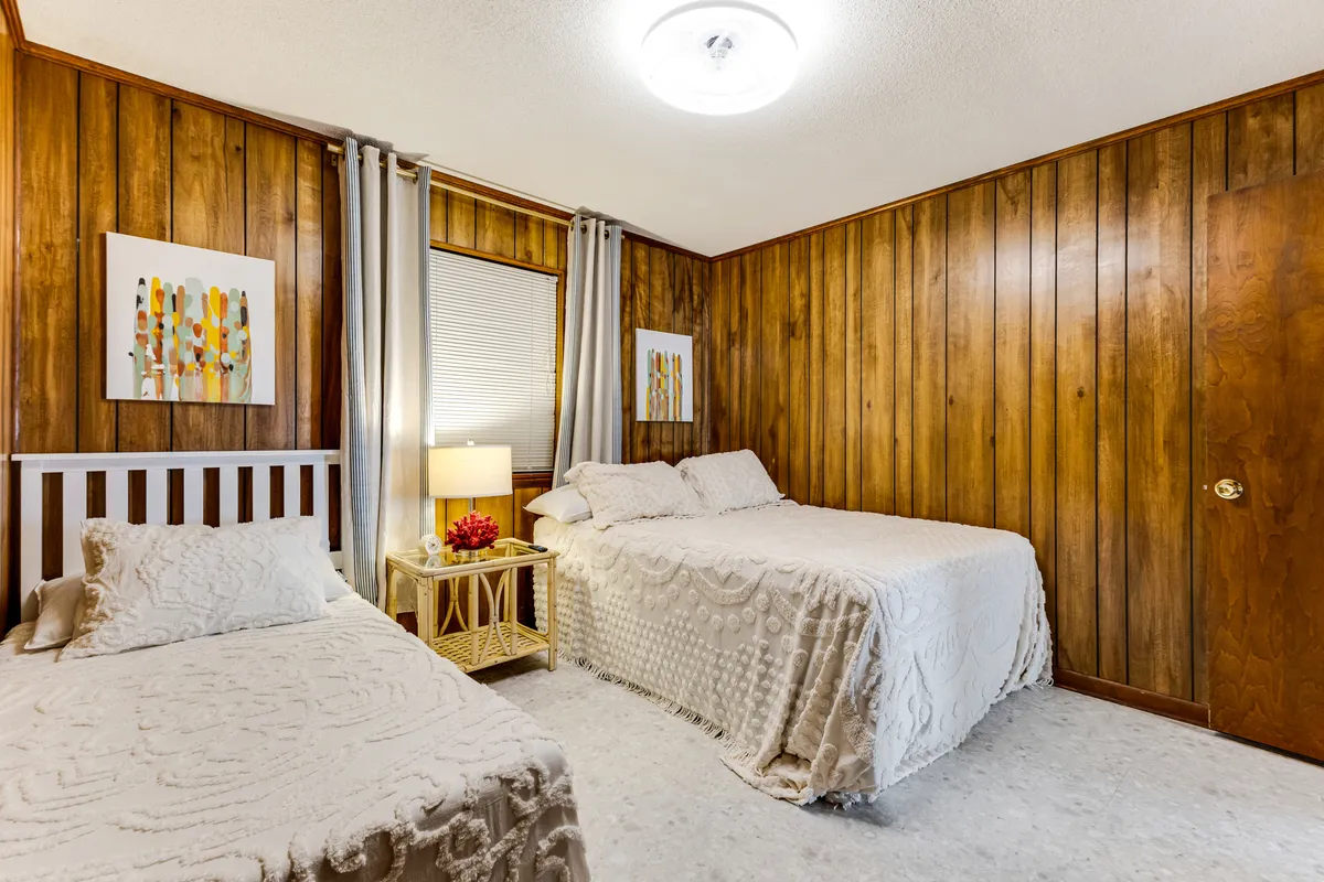 A cozy bedroom featuring two beds against warm wooden panel walls, each with soft bedding. A nightstand with a single flower sits between them, complemented by whimsical artwork, creating a relaxing and inviting retreat.
