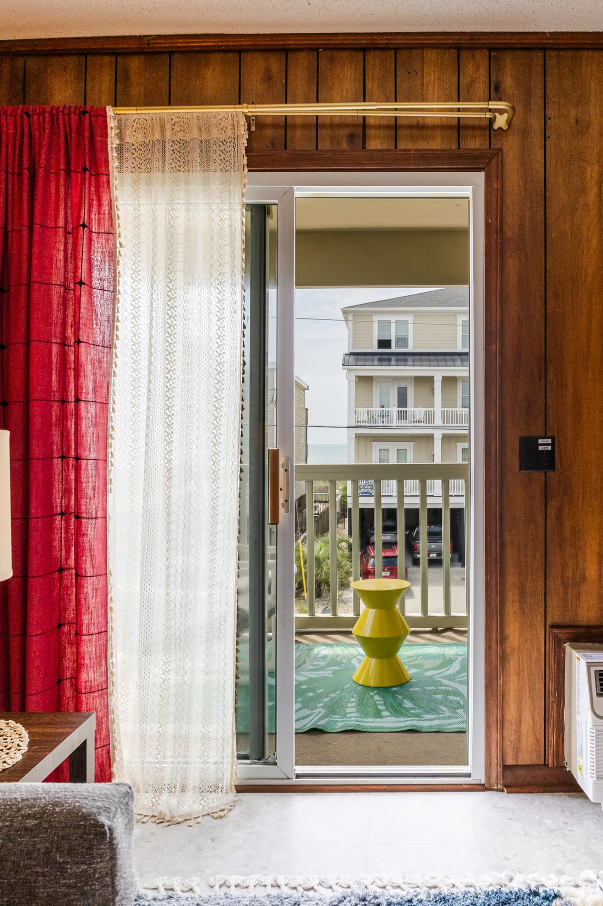 A bright interior space featuring a cheerful red curtain, a sheer white curtain, a yellow side table, and a clear view out to the balcony. The image hints at a seamless transition from indoor comfort to outdoor experiences, with natural light flooding the room and the balcony view inviting exploration.