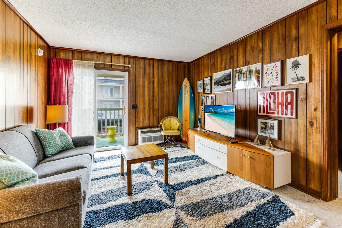 A funky, retro-inspired living area featuring a sofa, coffee table, television, and prominently displayed surfboard. The vibrant artwork and eclectic décor set a distinctly 1970s mood, inviting guests to relax or snap a selfie before heading out to explore.