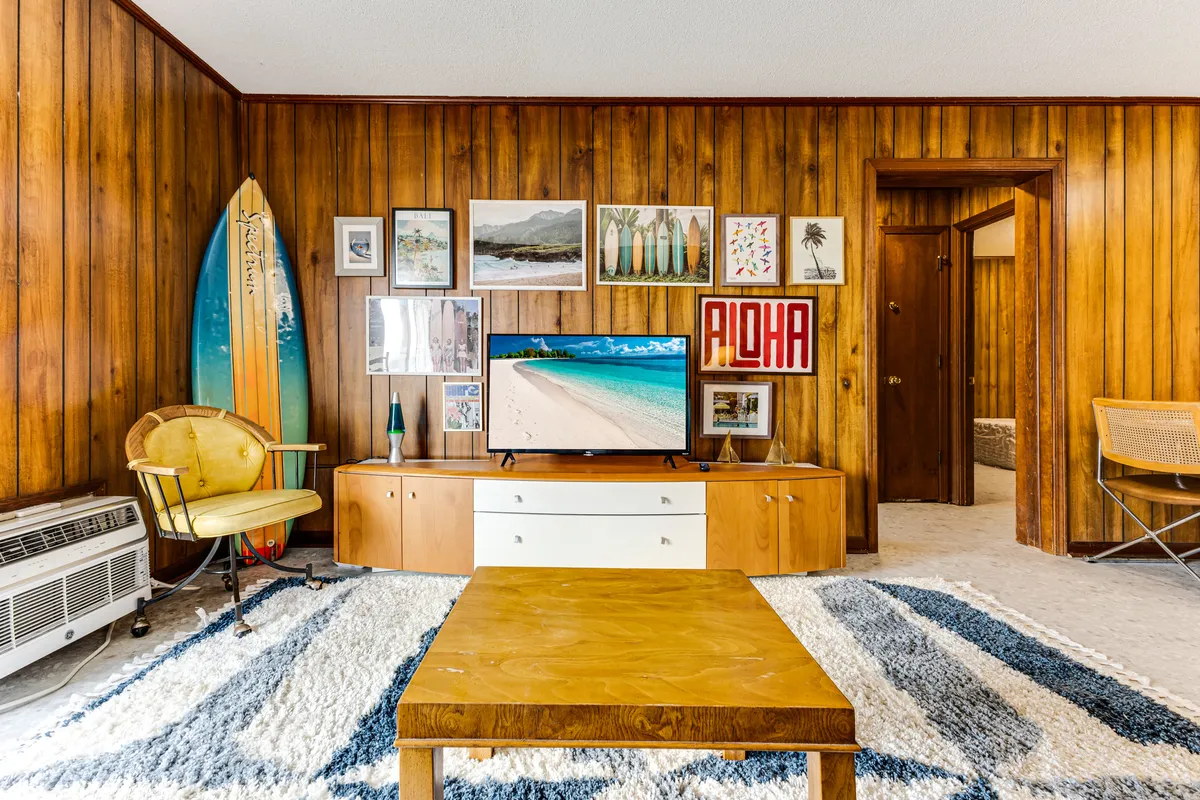 A bright and inviting living room decorated with retro flair, featuring a colorful surfboard, vibrant wall art, a cozy chair, and a modern TV. The space evokes a laid-back, beachy vibe perfect for unwinding after a day in the sun or gathering for game night with family and friends.