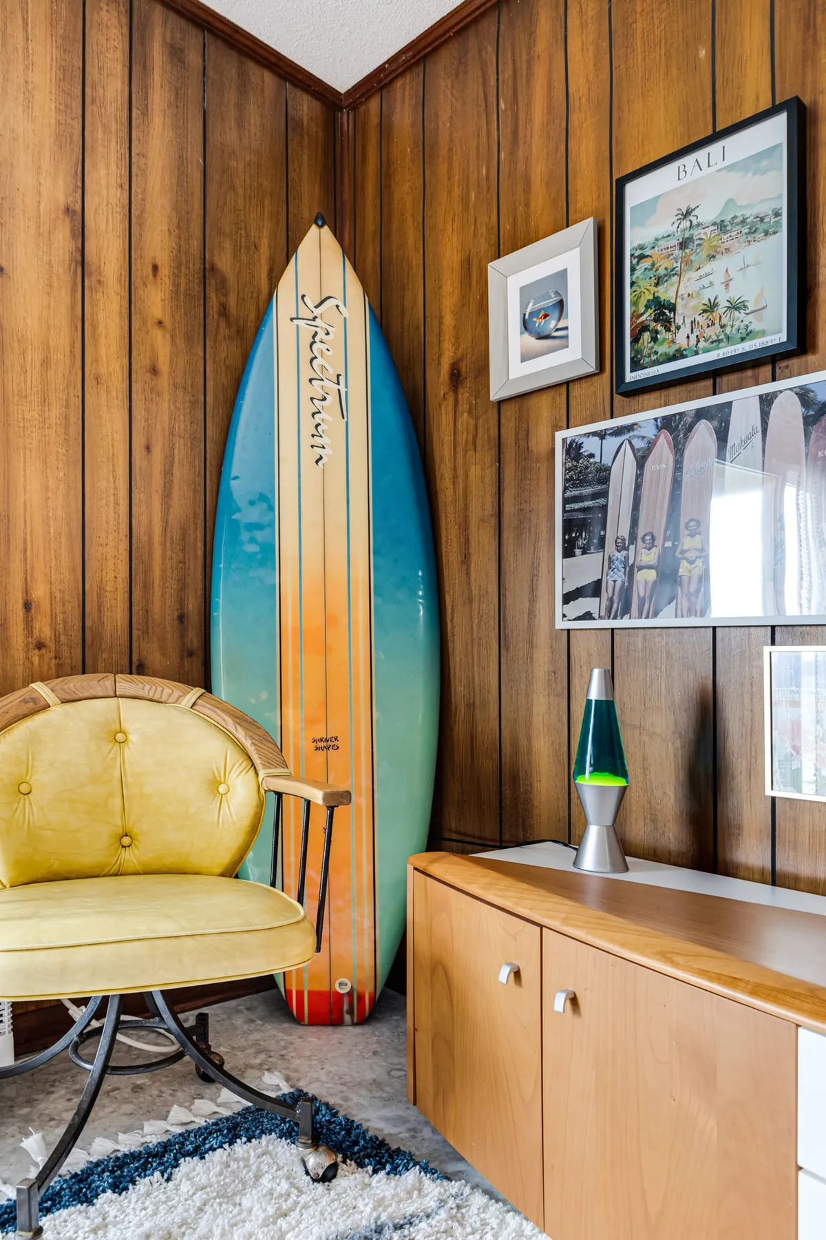 A playful, sunlit living area featuring a prominent surfboard, a cheerful yellow chair, a glowing lava lamp, and vibrant wall art that collectively evoke a 70s vibe. The scene feels energetic and nostalgic, with decor elements that nod to retro style and a relaxed, social atmosphere.