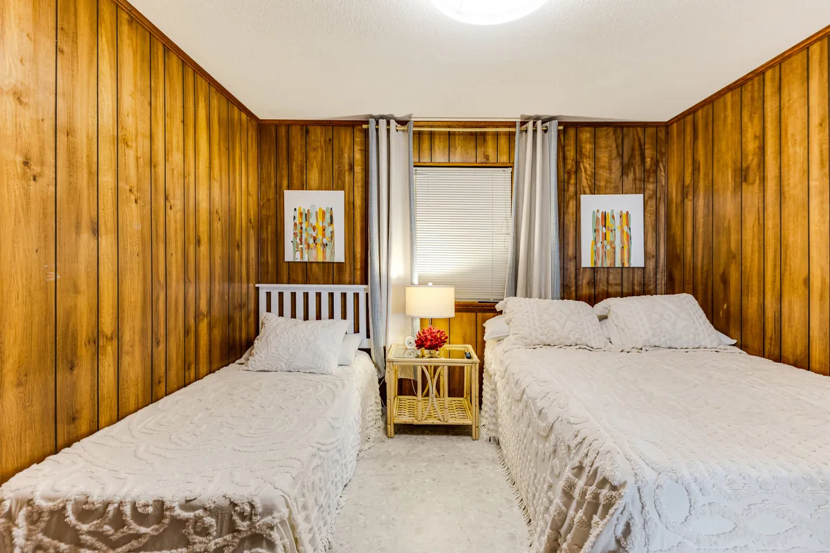A bright, inviting bedroom with two twin beds topped with crisp white bedding and playful accent pillows. A nightstand sits between the beds, adorned with a lamp and fresh flowers, while decorative retro-inspired wall art adds a burst of color and nostalgia.