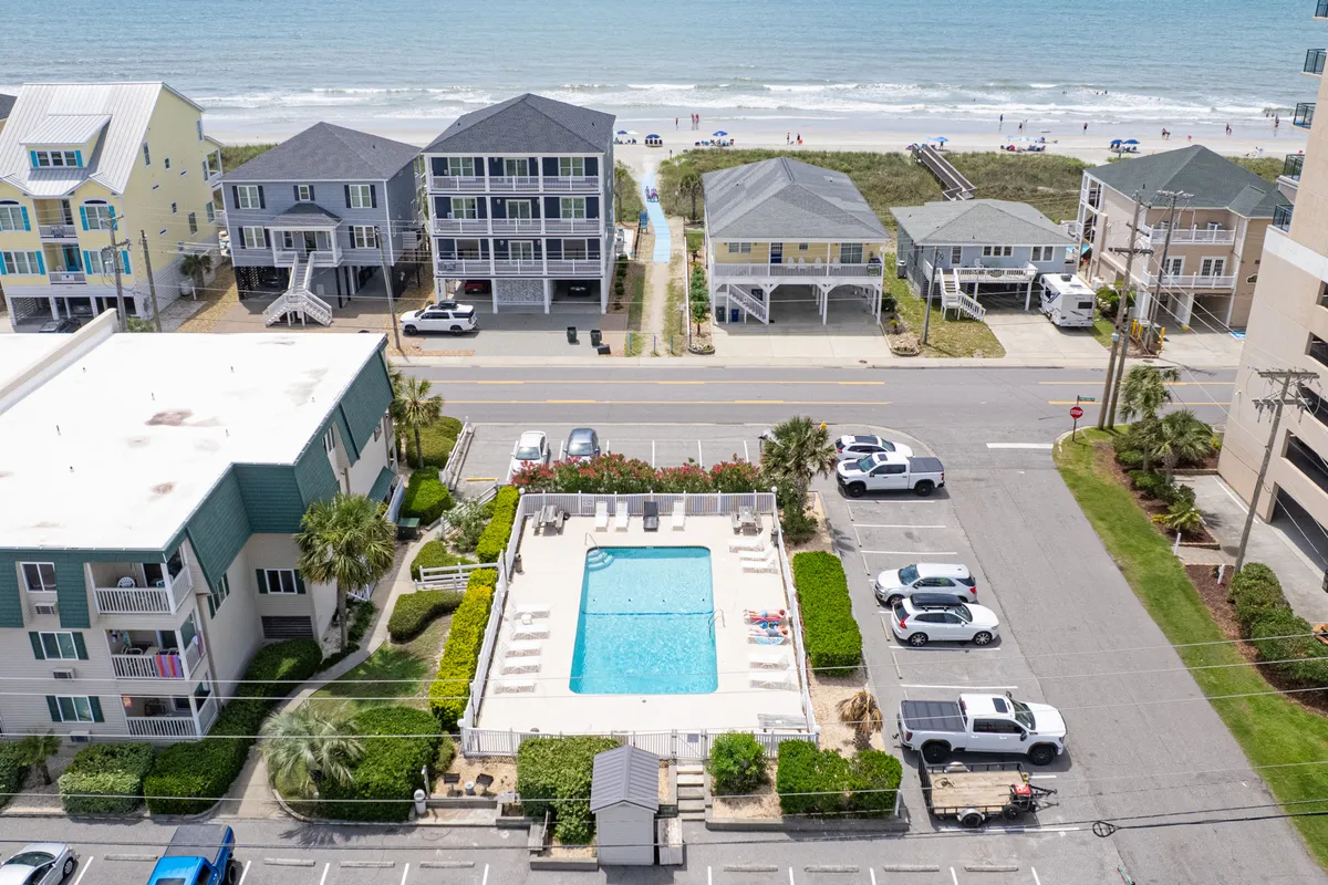This exterior image captures both a sparkling swimming pool and the ocean beyond, with a clear pathway leading directly from the pool area towards the beach. Surrounding houses and lush landscaping frame the view, while lounge chairs invite relaxation poolside. The image exudes a relaxed, vacation-ready mood with the amenities and location at the forefront.