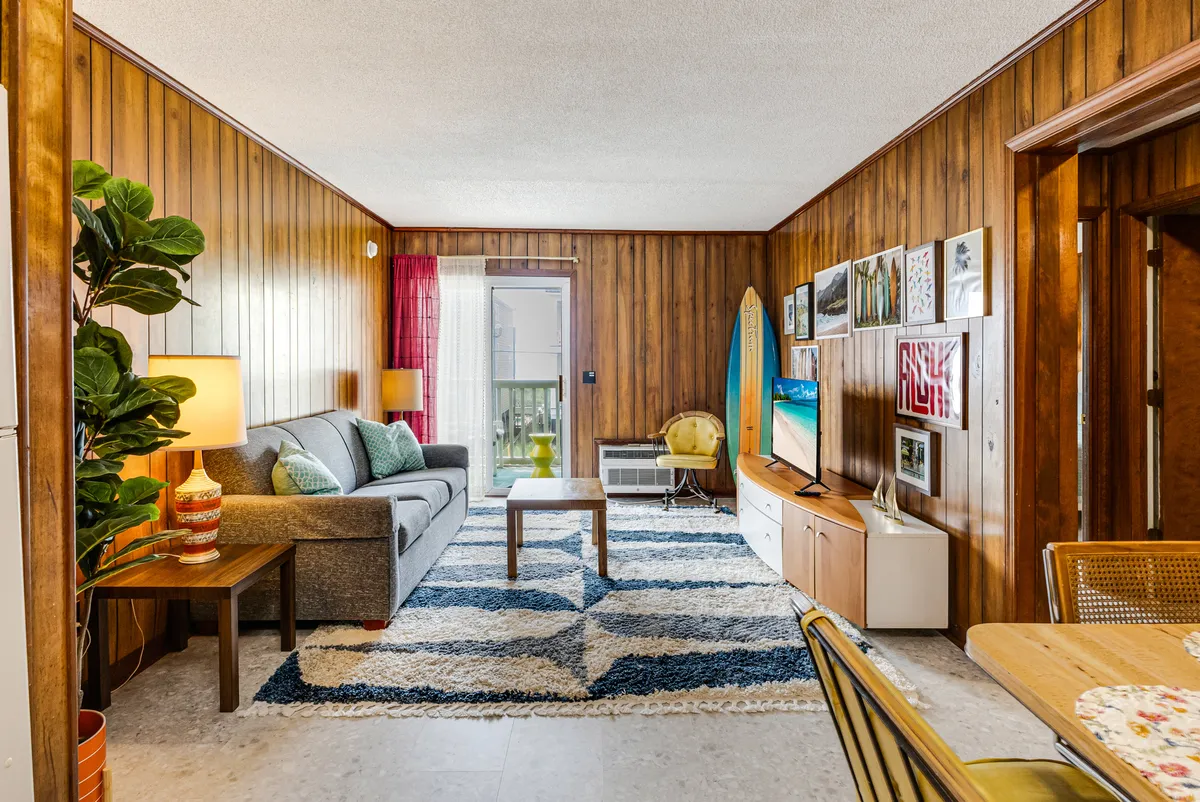 A comfortable living room featuring a retro-inspired sofa, a coffee table, a surfboard propped up as quirky décor, and colorful artwork adorning the walls. The space exudes a laid-back, stylish vibe perfect for relaxing after a day at the beach or socializing with friends.