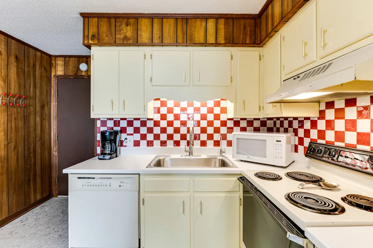 This image showcases a kitchenette with a lively checkerboard backsplash, crisp white cabinets, a stainless steel sink, and visible kitchen appliances. The retro touches in the backsplash and cabinetry, combined with modern conveniences, evoke a sense of nostalgia while highlighting practicality.
