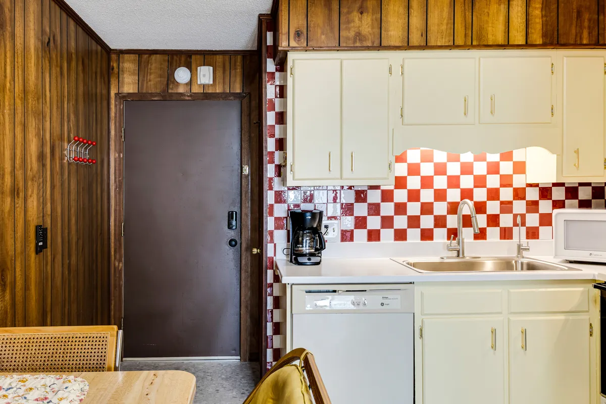 A broader view of the kitchenette reveals the red and white checkered backsplash, warm wood paneling, kitchen appliances, and a cozy dining table. The inviting mood is emphasized by the open, sociable layout, perfect for gathering friends for breakfast or snacks.