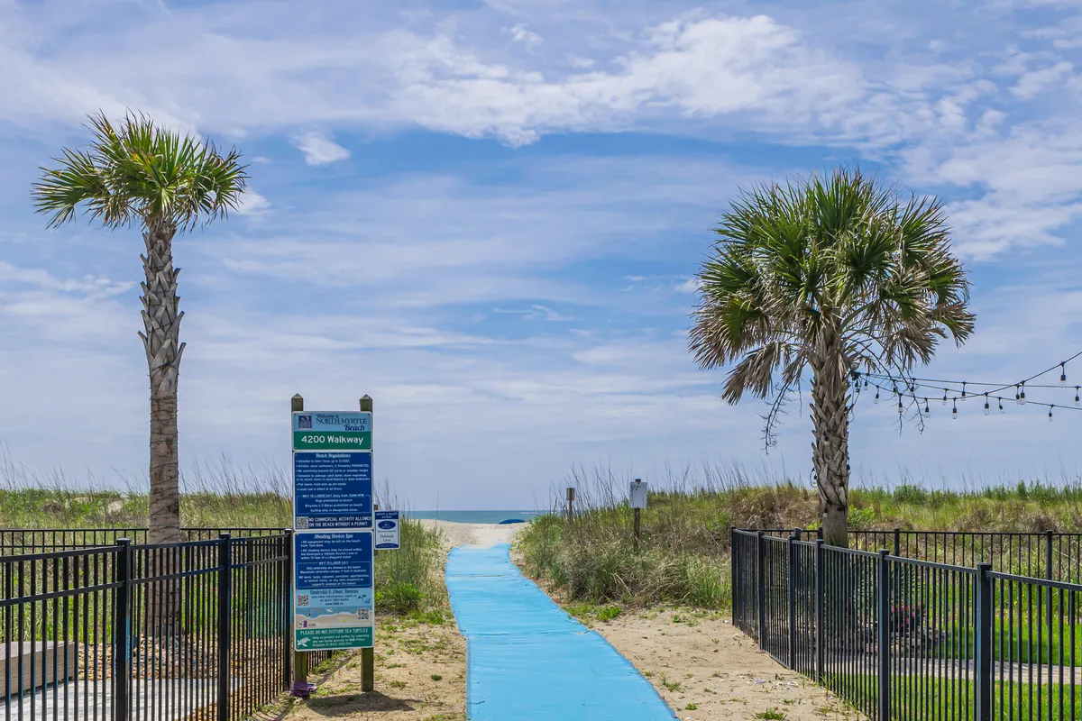 A tranquil exterior view of the blue walkway lined with palm trees, leading directly down to the sandy beach. The beach access sign warmly welcomes vacationers as sunlight brightens the path, hinting at the fun that lies just a few steps ahead.