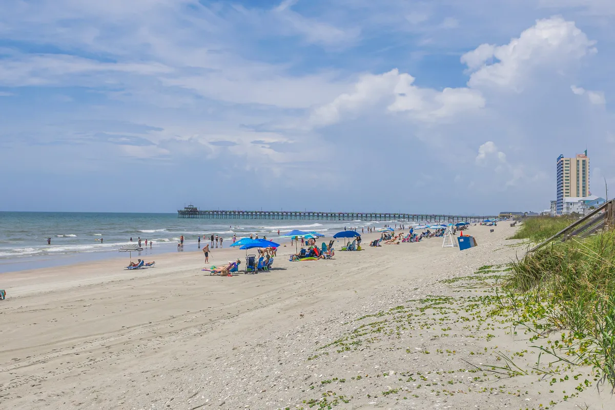 This vibrant exterior shot captures families and groups of people lounging under colorful beach umbrellas on the sandy shore, children playing and building sandcastles, swimmers enjoying the surf, and the iconic Cherry Grove Pier stretching into the ocean in the background. Distant buildings anchor the scene as a lively, family-friendly beach destination.